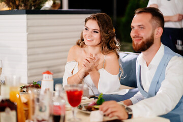 newlyweds sit at table in restaurant and listen to congratulations from guests.
