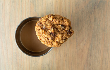 Top view of a single homemade oatmeal raisin cookie on the rim of a coffee cup atop a table illuminated with natural light.
