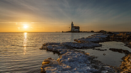 Fototapeta na wymiar Winter landscape at the Lighthouse Paard van Marken Netherlands. On a cold day, temperature -7 degree during sunset. The IJsselmeer is frozen and by strong wind the shore was covered with ice