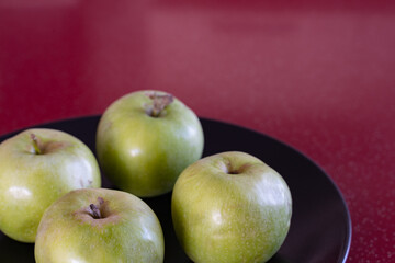 A few green fresh apples lie on a black platter on the red table.