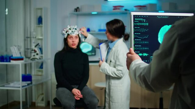 Stressed patient sitting on neurological chair with eeg headset, testing health status of brain, while medical researcher looking at monitor with tomography scan developing treatment in laboratory