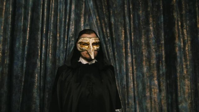 lonely mystical man actor in a black robe and with a hood on his head wearing a Venetian mask opens the curtain with his hands and enters the stage