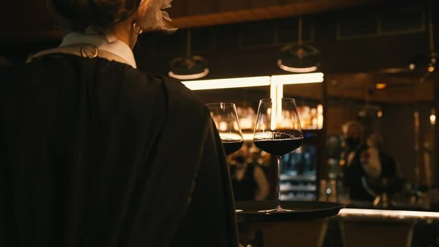 a girl waitress in a Venetian mask with a white feather and in a black robe walks with a tray of glasses of wine through a luxury restaurant and leaves the wine room