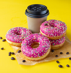 Yummy pink glaze donuts with colorful sprinkles, takeaway coffee cup and roasted coffee beans on yellow background. Junk food.