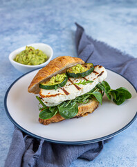 Croissant sandwich with grilled halloumi cheese, arugula and grilled zucchini, blue concrete background. Healthy breakfast or lunch.