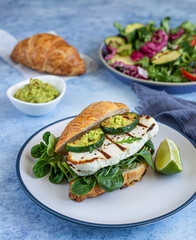 Croissant sandwich with grilled halloumi cheese, arugula and grilled zucchini, blue concrete background. Healthy breakfast or lunch.