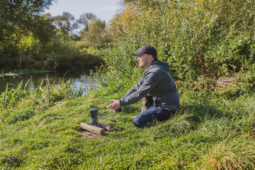 Man sits alone with a cup of coffee on the river bank near the fire. Rest at nature. Man sits alone near a campfire by the river on a sunny day.
