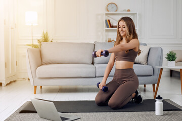 Fototapeta na wymiar The young beautiful sports girl in leggings and a top does exercises with dumbells. Healthy lifestyle. A woman goes in for sports at home while using laptop.