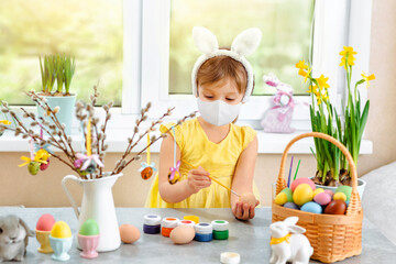 A girl in a medical mask paints eggs for Easter holiday in different colors with a brush.