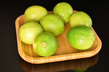 Several bright green juicy limes on a wooden dish, close-up, on a black background.
