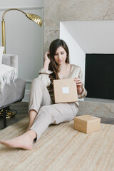 
A beautiful dark-haired girl is sitting in the living room against the background of the fireplace with a box in her hands. The concept of comfort, style.