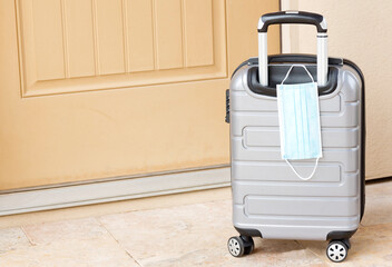 Pandemic travel seen in tan door, gray suitcase, and face mask reflects business and leisure travel within coronavirus public health guidelines