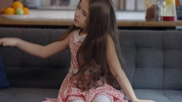 A beautiful little girl with long hair is sitting on an armchair in her home, dancing and playing with her hair. Portrait of beautiful little girl with long hair. Slow motion video.