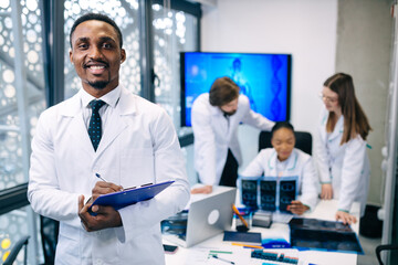Fototapeta premium Smiling doctor, holding a notebook with notes, stand in front, behind his back the team is discussing the patient's X-ray