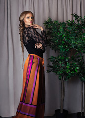 Portrait of a beautiful white woman with curly long blond hair and beautiful makeup posing by herself in a studio with grey curtain background wearing a sheer black blouse and colorful pants.