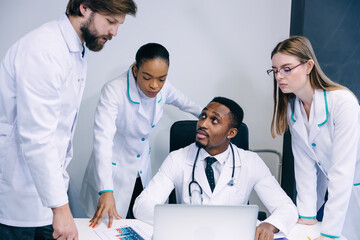 hospital, profession, people, technology and medicine concept - group of doctors with clipboard and laptop computer meeting and discussing something at medical office