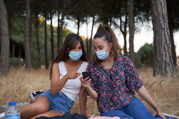 Two friends look at the cell phone while one of them points at it in surprise while they are having a picnic in the forest protected by a mask
