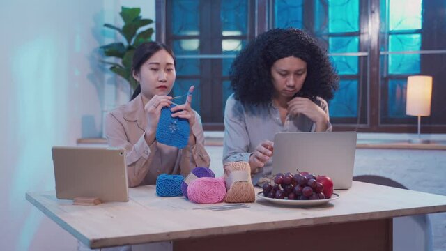 Hipster business male freelance using laptop working at home office while female doing hobby by crochet handcraft in living room at night. Young Asian Couple spend time together during lockdown