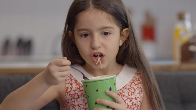 Little girl eats noodles from a glass at her home. Ready and fast food concept. Creative people can change the track-point package in the girl's hand.