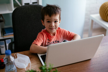 Portrait of cute smiling little boy sitting on office chair behind desk in the office. Happy boy using laptop.