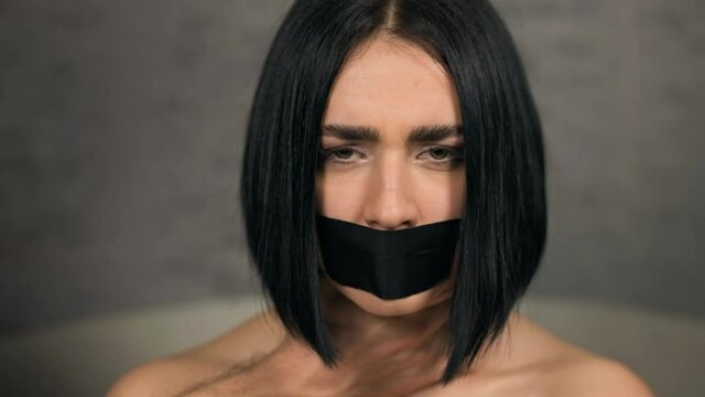 Serious woman with duct tapered mouth grabbed by male hands over neck looking at you