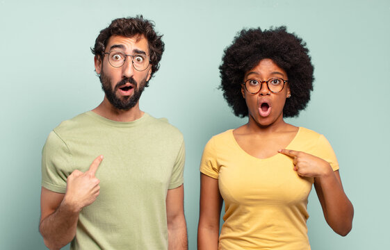 multiracial couple of friends looking shocked and surprised with mouth wide open, pointing to self