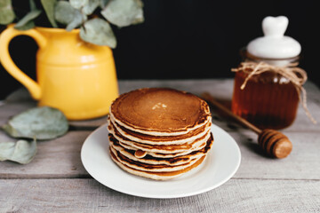 Pancakes with honey on plate, wooden table, spoon, yellow vase with eucalyptus
