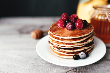 Juicy pancakes with berries and honey on a white plate, spoon, jar, wooden table