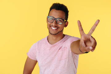 Smiling successful african american young man showing peace gesture over yellow background.