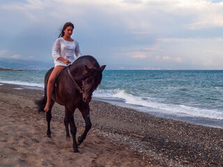 Joven amazona con vestido blanco domando a su caballo marron sin montura en la playa