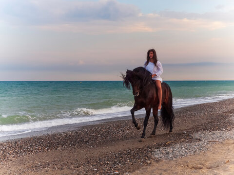 Joven amazona con vestido blanco domando a su caballo marron sin montura en la playa