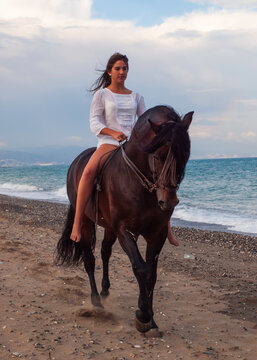 Joven amazona con vestido blanco domando a su caballo marron sin montura en la playa