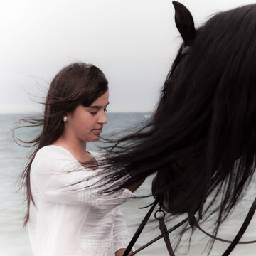 Joven amazona con vestido blanco domando a su caballo marron sin montura en la playa