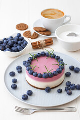 The two-color blueberry mini cheese cake. Round no bake cheesecake, cut portion on small plate with few cookies in the focus. The top of cake decorated by rosemary herbs. White table.