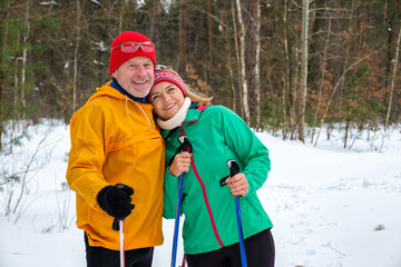 Senior couple resting while walking with nordic walking poles. Smiling, hugs and looking at the camera in a winter park. Elderly wife and husband go in for sports in nature. Active lifestyle concept.