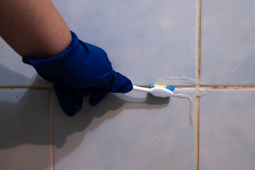 A hand in a blue glove cleans the dirty joints between the tiles in the bathroom with an old toothbrush. Toxic black mold in the bathroom during the cleaning process.