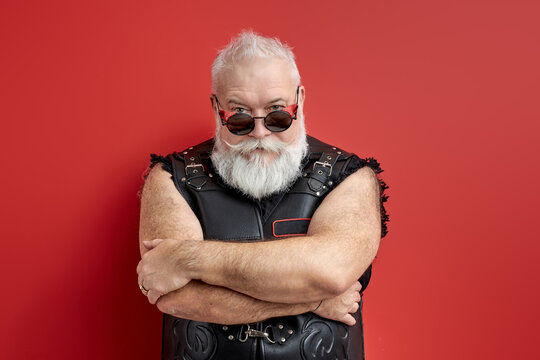 biker in leather jacket looking at camera from under the eyeglasses, isolated on red studio background. Cool gray bearded male confidently posing, standing with crossed arms