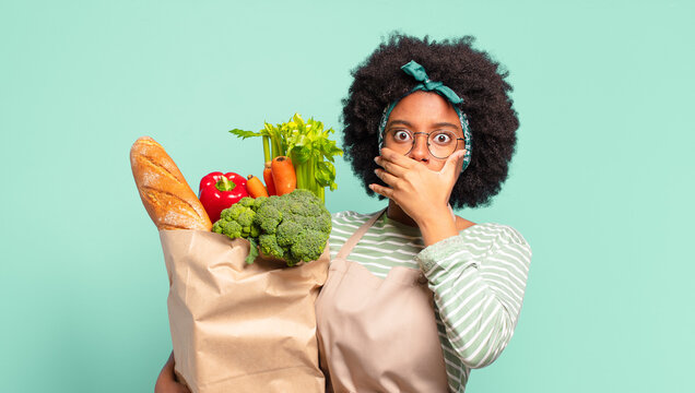 young pretty afro woman covering mouth with hands with a shocked, surprised expression, keeping a secret or saying oops and holding a vegetables bag