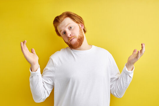 Redhead man shrugging, wearing casual sweater over isolated yellow background clueless and confused expression with arms and hands raised. Doubt concept.