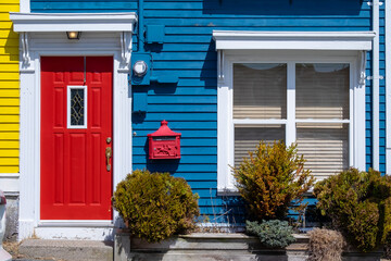 Fototapeta na wymiar A bright red metal door of a building with blue and yellow wooden clapboard walls. The house has a red decorative metal mailbox. There's a green shrub near the red door with white trim. 
