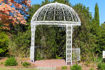 A gorgeous shot of a white rod iron gazebo surrounded by lush green grass, trees and plants near a pink and yellow tree at South Coast Botanic Garden in Palos Verdes, California - Powered by Adobe