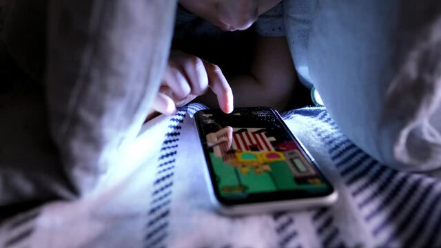 kid, a boy of 8 years old lies in bed under the covers, plays in the application on a smartphone, the concept of addiction, insomnia