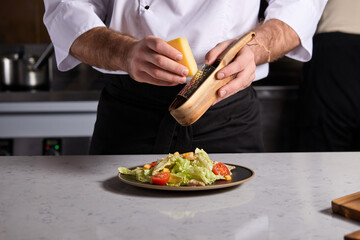 cropped chef chop cheese on grater, making fresh salad in restaurant kitchen, wearing uniform. Male professional prepare dish for guests
