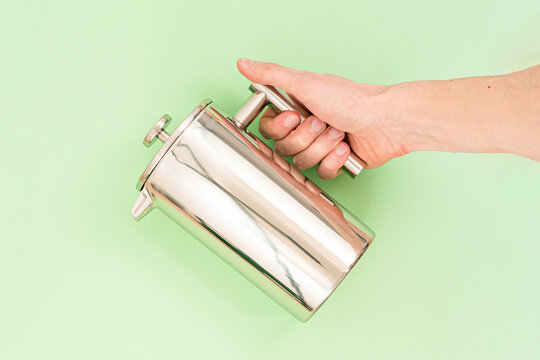 Man's hand holds stainless steel french press on a light green background. Filter. Design. Kitchen. Tea. Coffee. Handle. Preparation. Useful. Device