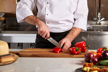 cropped chef man cutting fresh red bell pepper using knife on wooden cutting board. young man in uniform cutting pepper for vegetable salad. concept of healthy eating.