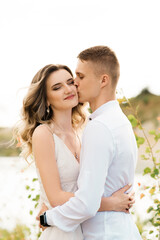 Fototapeta premium A beautiful young couple in love, a man and a woman embrace, kiss near a blue lake and sand at sunset. Vacation at the sea on the beach, sunset, morning, honeymoon, tropics, ocean, girl, relationship.