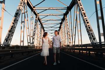 A beautiful young couple in love, a man and a woman, embrace, kiss on a large metal bridge at sunset.