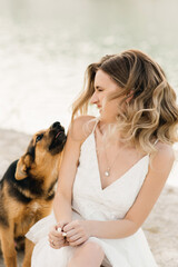 Portrait of a beautiful girl in a white dress with hair and makeup playing with a dog on the background of the sea