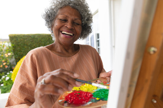 African american senior woman smiling while painting on canvas standing on porch of the house