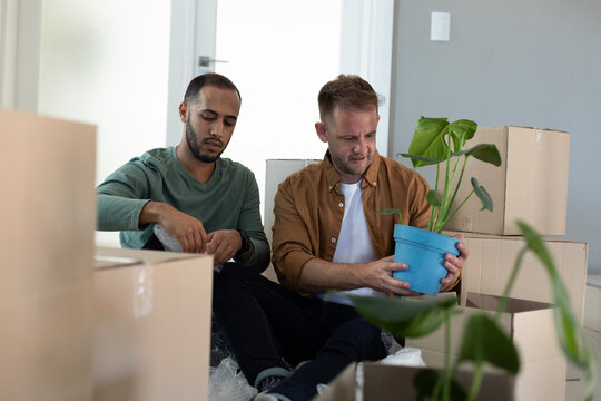 Multi ethnic gay male couple sitting surrounded by boxes and holding plants at home